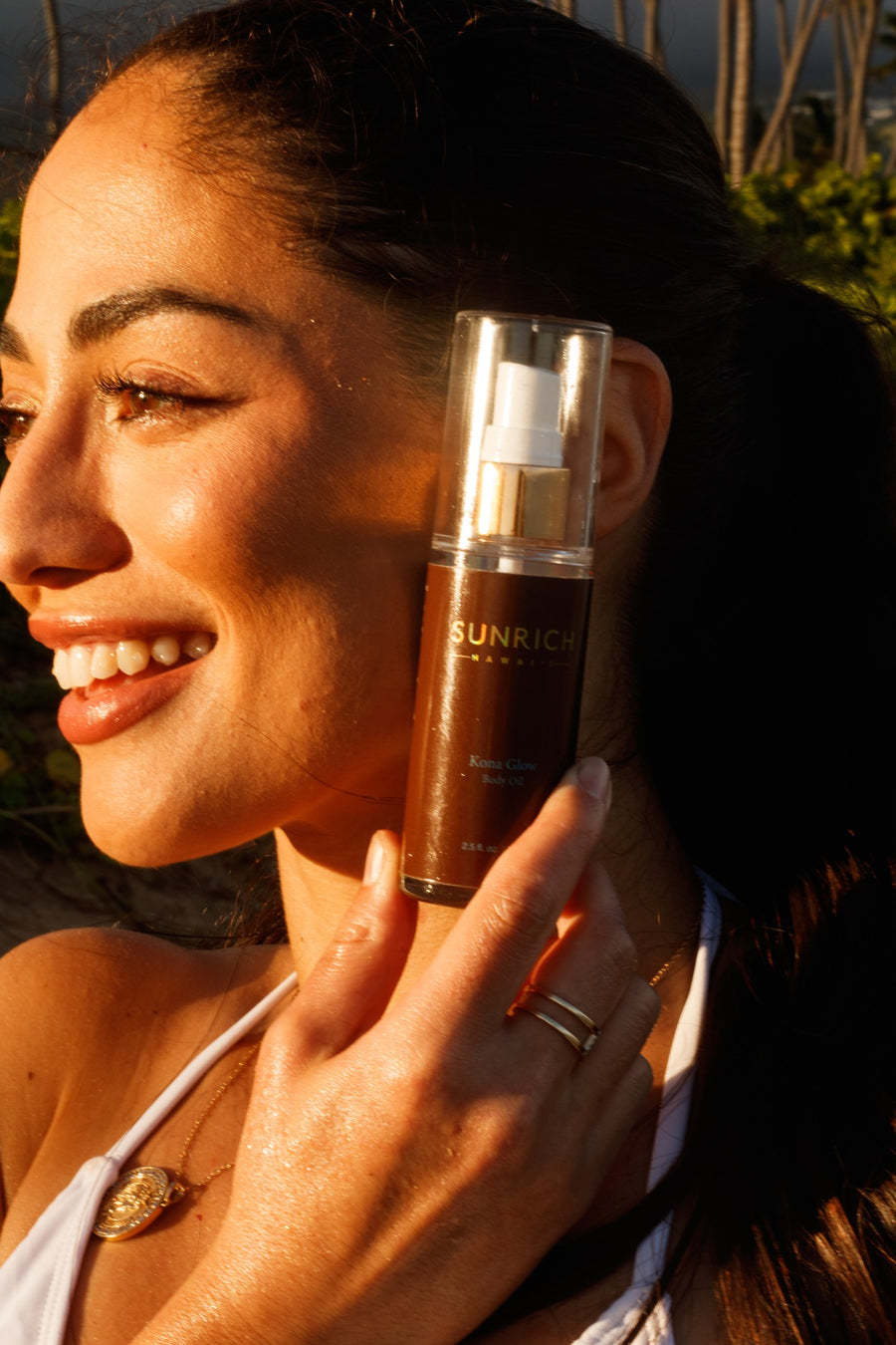 A woman in golden-hour light holding a bottle of SUNRICH Kona Glow Body Oil to her cheek, skin softly luminous
