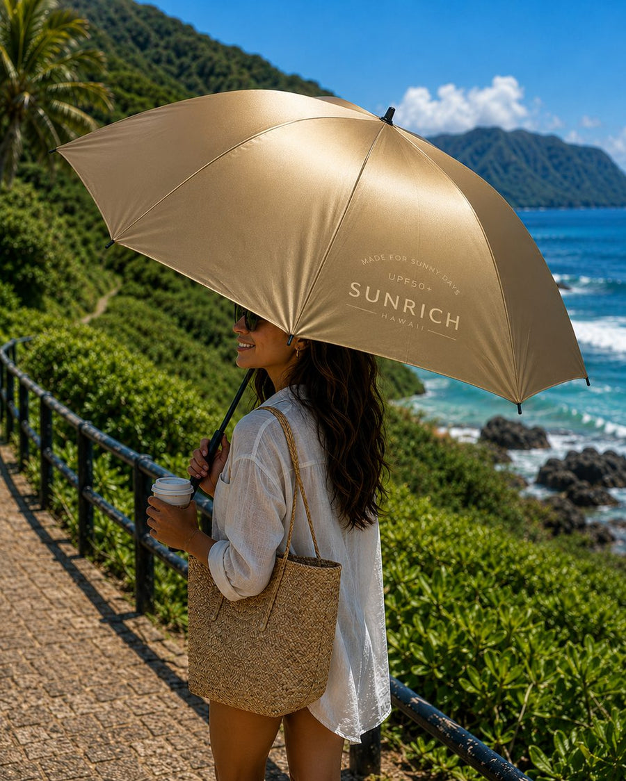 A woman walks a coastal path holding the metallic gold SUNRICH Sun Shade open above her, with green cliffs and ocean behind