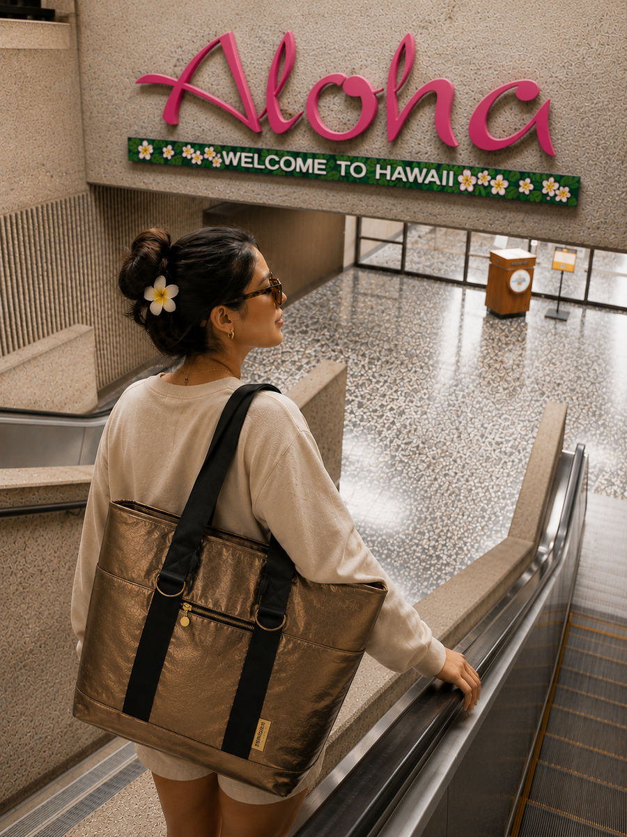 A traveller rides an escalator beneath an Aloha Welcome to Hawaii airport sign with the SUNRICH Sun Tote in gold on her shoulder