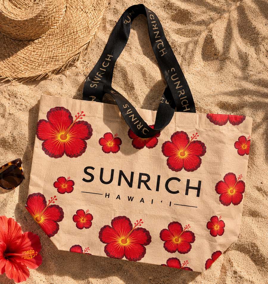 The SUNRICH Hibiscus Sun Market Tote in natural canvas with a bold red hibiscus print and black branded straps, on sand beside a straw hat and sunglasses